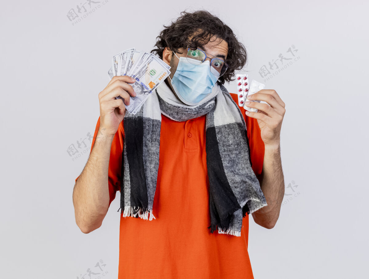 年轻印象深刻的年轻白种人病人戴着眼镜 围巾和口罩拿着钱和药丸看着隔离在白色背景上的相机钱持有眼镜