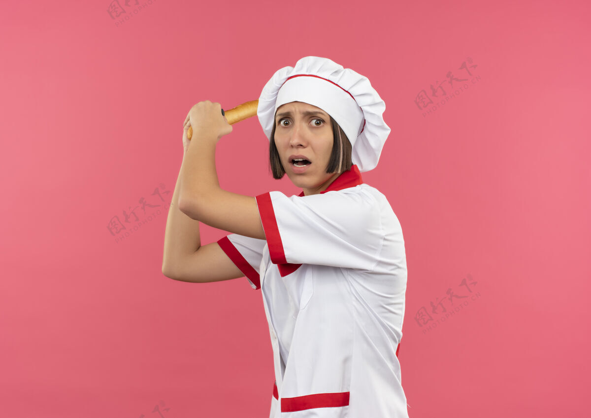 有人愤怒的年轻女厨师穿着厨师制服 拿着面包棒 准备用复印空间打一个孤立的粉红色的人女性年轻复制