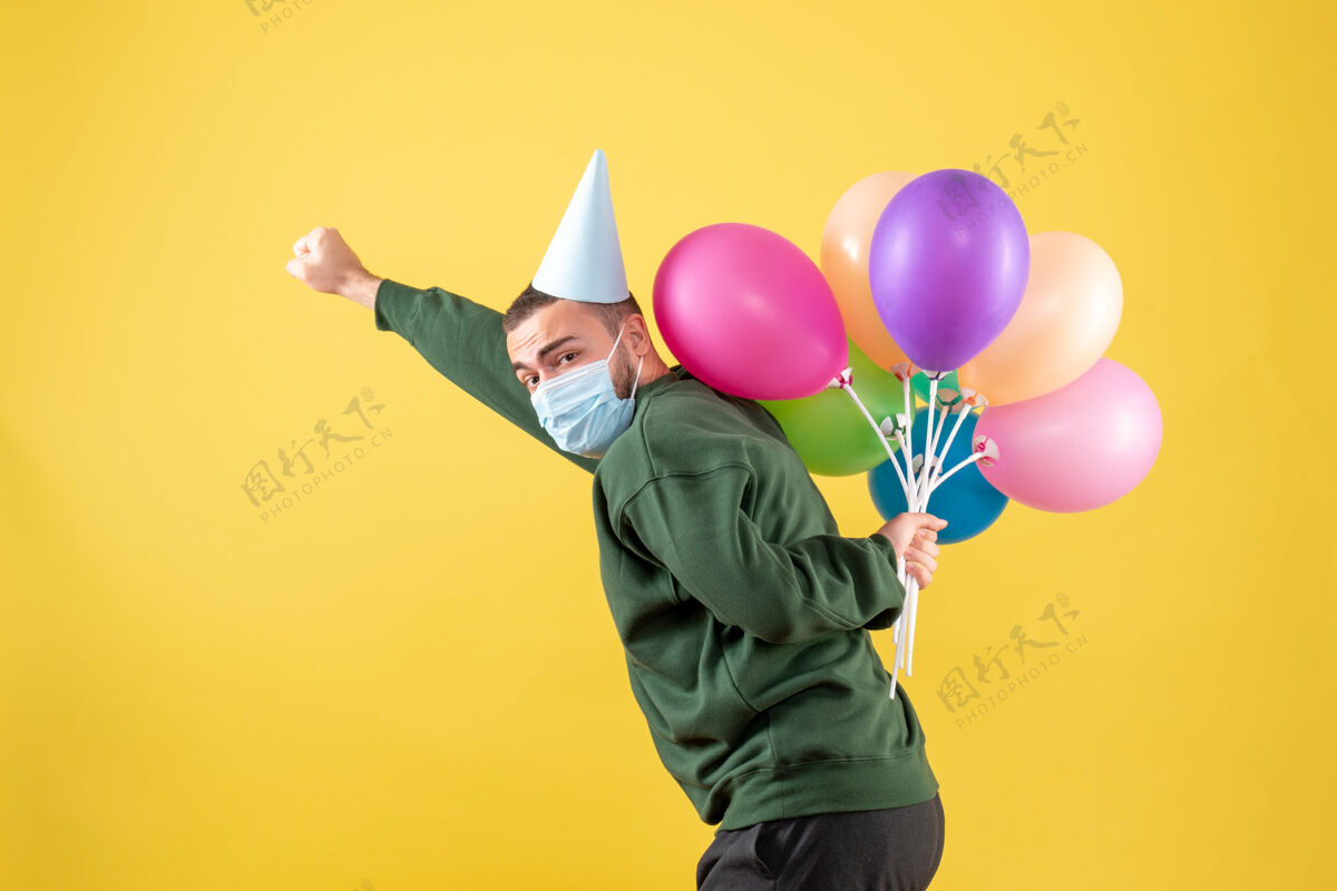 生日正面图：黄色背景上戴着面具手持彩色气球的年轻男性大流行气球聚会