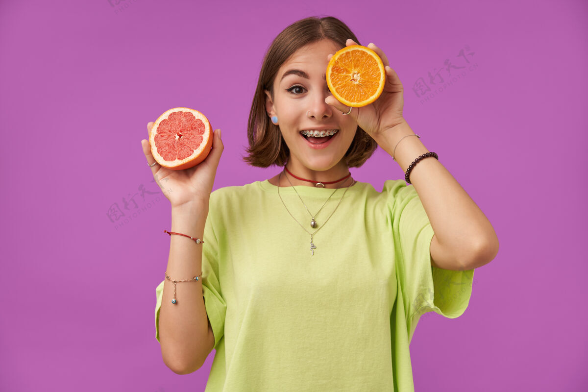 休闲女学生 年轻的惊讶女士 深色短发 用橙色遮住眼睛 遮住一只眼睛 站在紫色的墙上 穿着绿色t恤 项链 背带和手镯微笑项链模特
