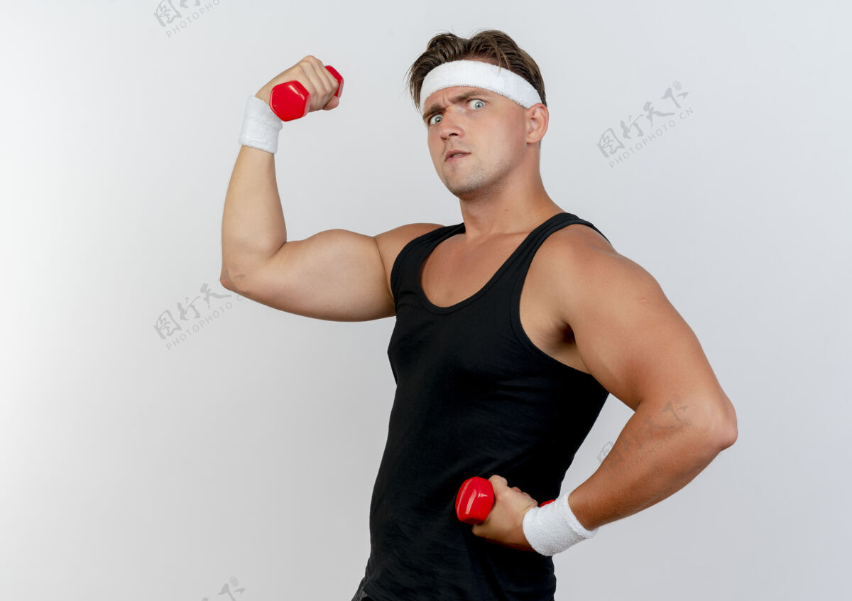 抱自信的年轻帅气的运动型男人戴着头带和腕带站在侧视图中举着哑铃在白色上做着强烈的孤立的手势看自信运动