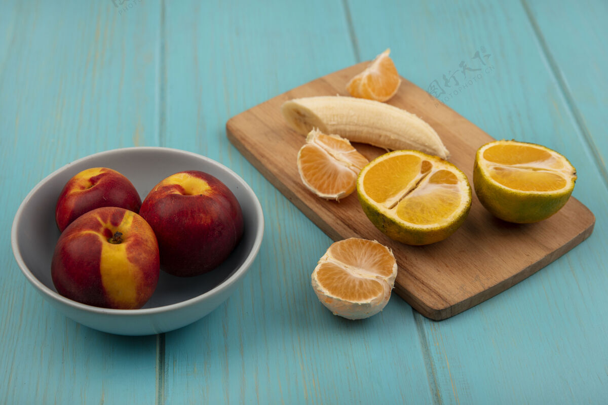 持有新鲜剥皮的香蕉放在木制厨房板上 橘子和桃子放在蓝色木墙上的桶上橘子手桶