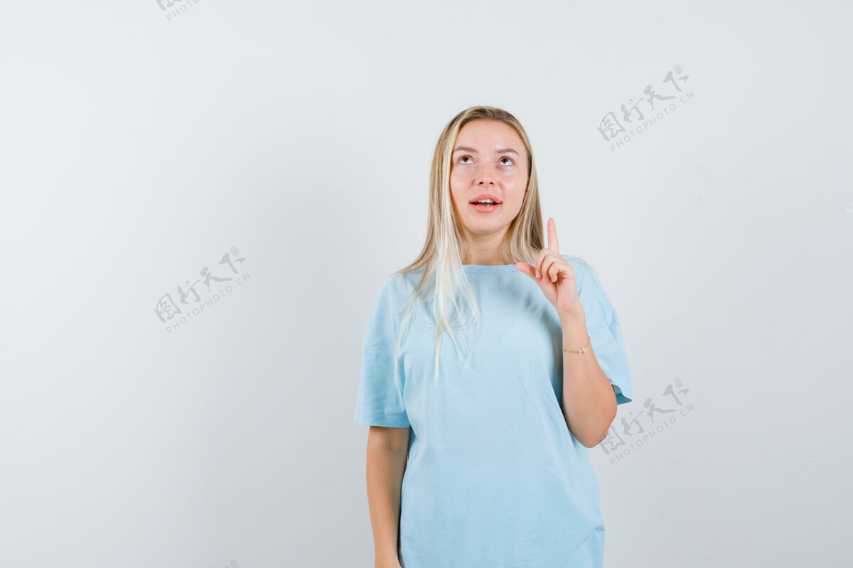 前面穿着蓝色t恤的金发女孩 食指朝上 向上看 注意力集中 正面视图时尚手指焦点