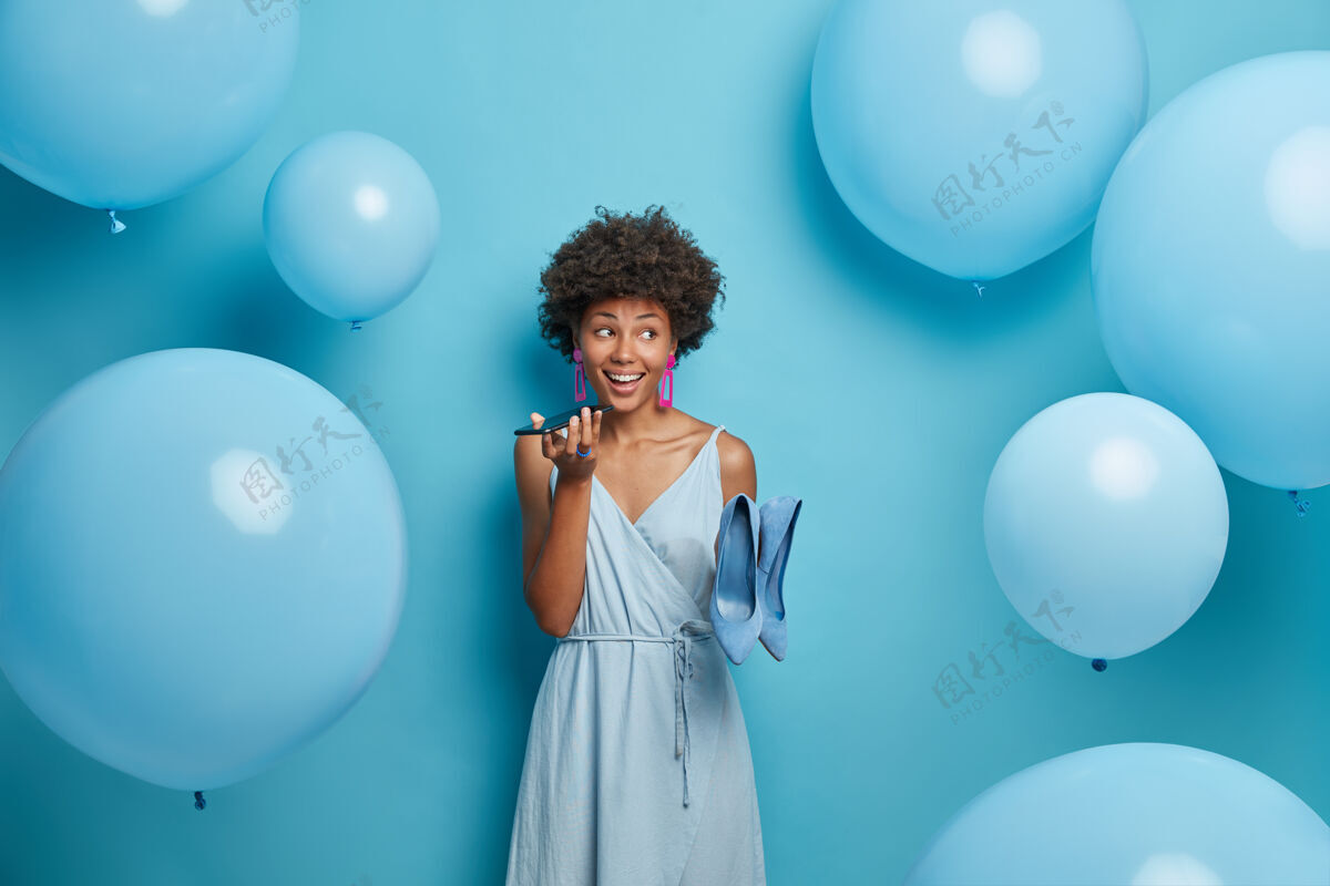 高跟鞋时尚的非洲裔美国妇女把手机放在嘴边 打电话 穿着时髦的裙子和高跟鞋 穿着节日活动的衣服 站在室内充气的氦气球旁边黑发女士魅力