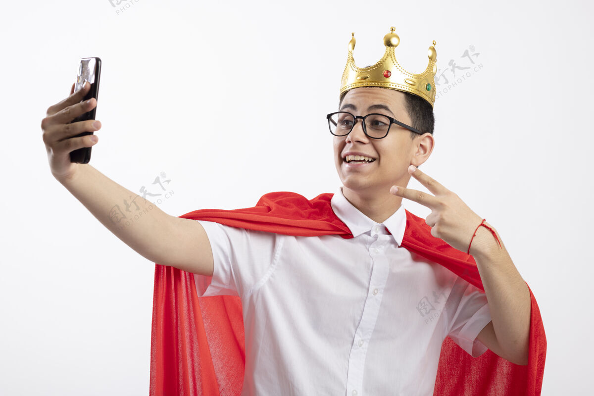白色快乐的年轻超级英雄男孩在红斗篷戴着眼镜和王冠做和平标志采取自拍隔离在白色背景上穿眼镜欢乐