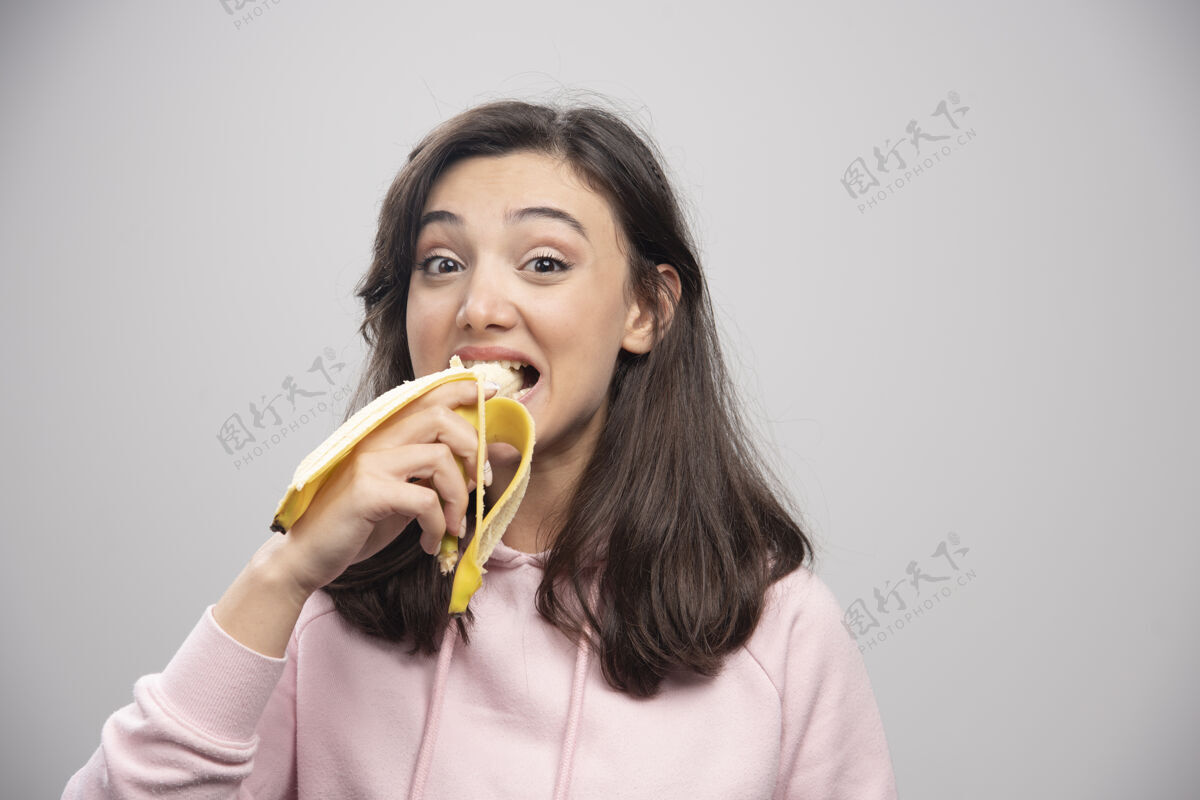 积极年轻的女人在灰色的墙上吃香蕉饮食女人微笑