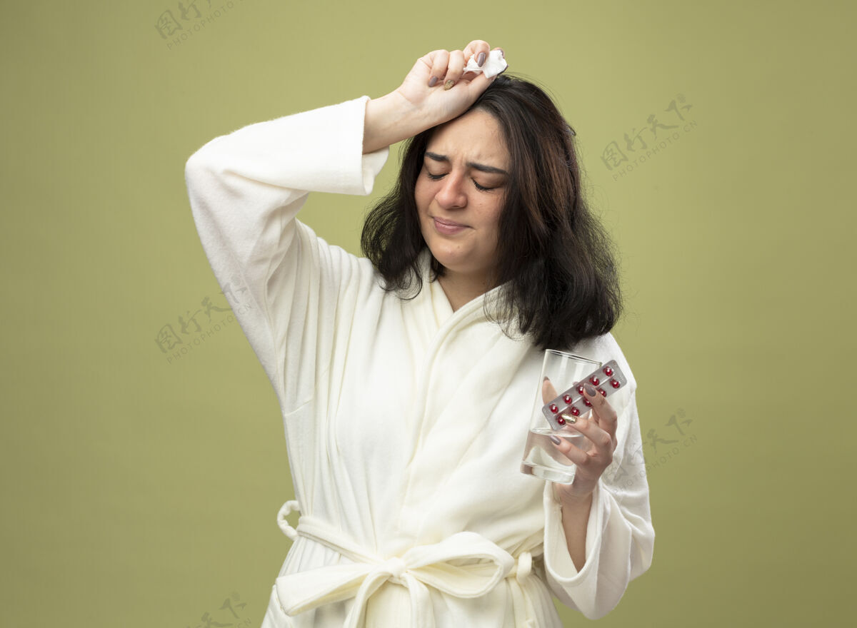 封闭疼痛的年轻白种人女孩穿着长袍拿着一包药丸一杯水和一张餐巾 闭着眼睛 在橄榄绿的背景上与复印空间隔离持有空间复制