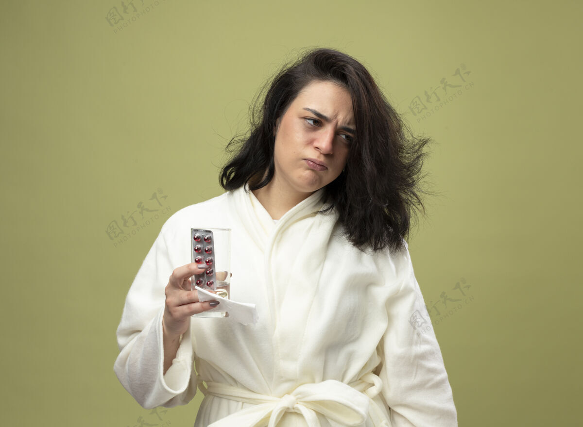 疾病愁眉不展的年轻白种女孩穿着长袍 手里拿着一包药丸 一杯水和餐巾纸 看着橄榄绿背景上孤立的一面包装年轻长袍
