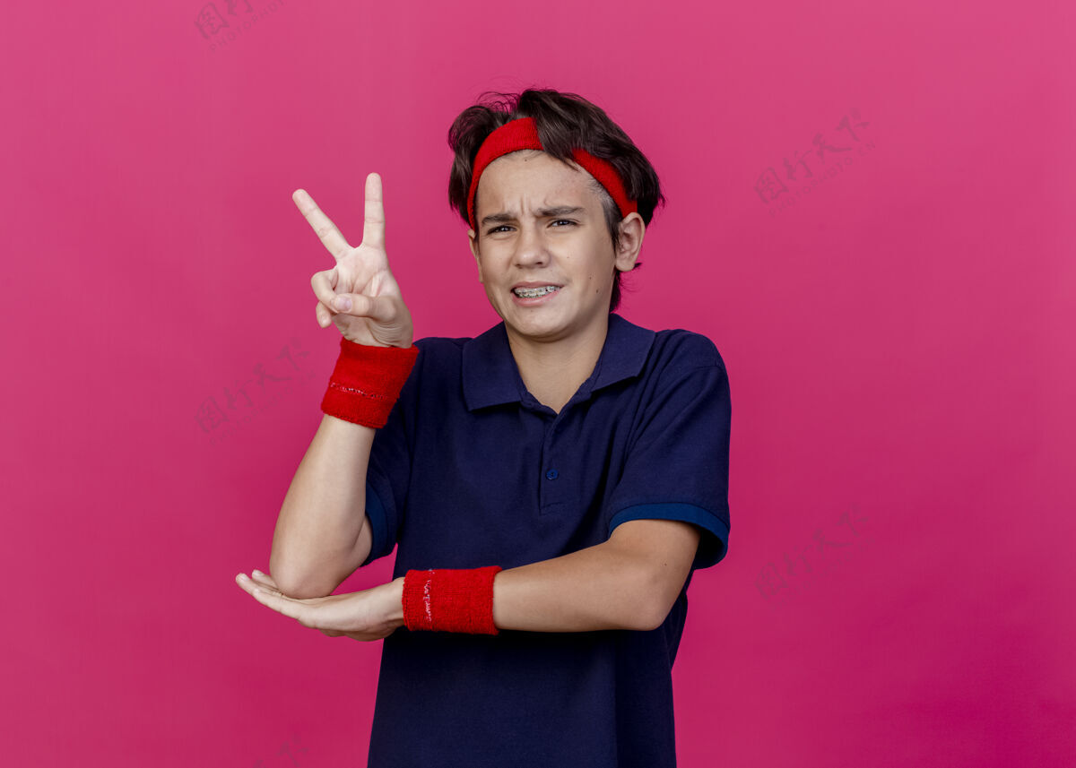 人皱眉的年轻英俊的运动男孩戴着头带和带牙套的腕带把手放在手肘下看着前面做和平的标志隔离在深红色的墙上表情人和平