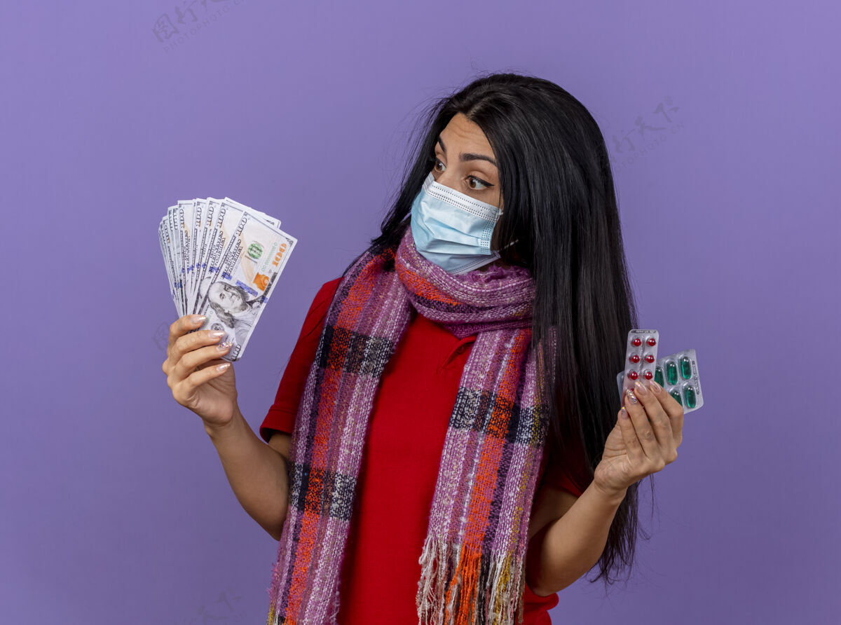 人印象深刻的年轻生病的女人戴着面具和围巾拿着钱和一包胶囊看着隔离在紫色墙上的钱站围巾钱