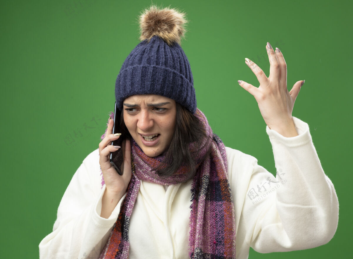 人愤怒的年轻生病的女人穿着长袍 戴着冬天的帽子和围巾在打电话 手举在空中 看着隔离在绿色墙上的一面公民保持脸