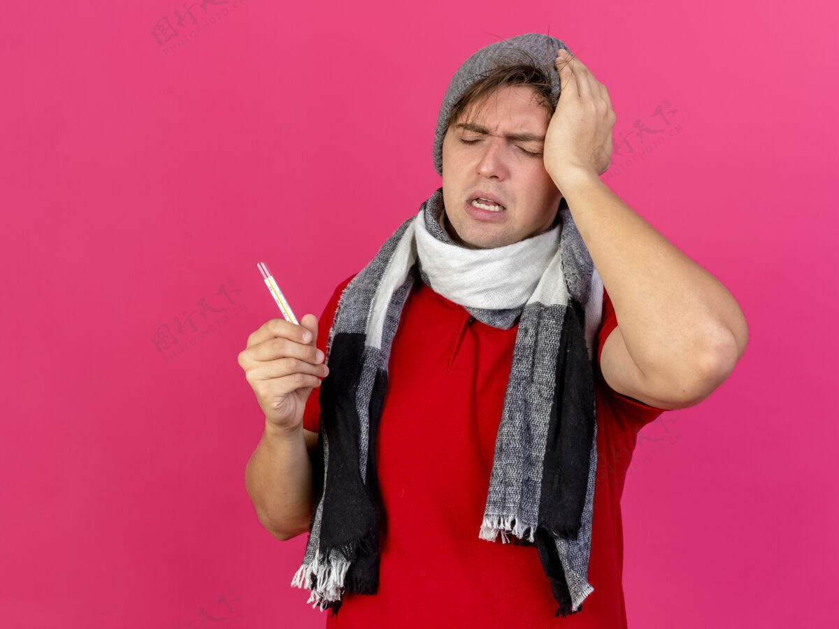 触摸疼痛的年轻英俊的金发病男子戴着冬天的帽子和围巾拿着温度计触摸着头闭着眼睛隔离在粉红色的墙上疼痛表情姿势