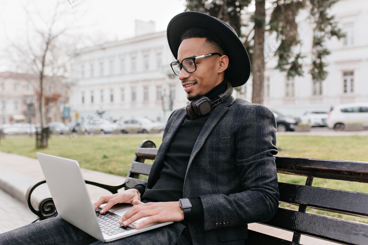 企业家帅气的年轻自由职业者在公园里用电脑工作一个戴帽子的快乐的非洲男人在长凳上用笔记本电脑学习的户外肖像成年人长凳公园