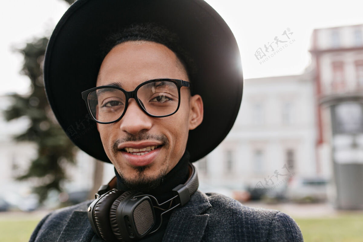 肖像戴着大帽子微笑的非洲年轻人的户外照片戴着黑色耳机的令人毛骨悚然的家伙在公园里消磨时光的特写照片男人音乐寒冷