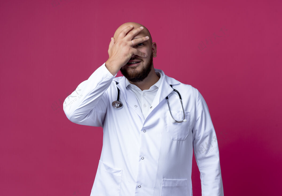 粉红色高兴的年轻男医生穿着医用长袍和听诊器捂着脸脸手男性