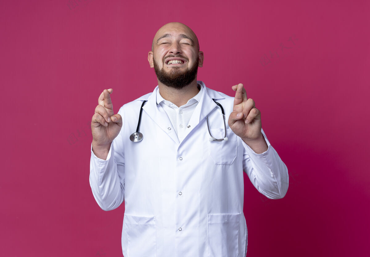医生年轻秃顶的男医生戴着医用长袍和听诊器闭着眼睛 手指交叉着隔离在粉红色上眼睛听诊器长袍