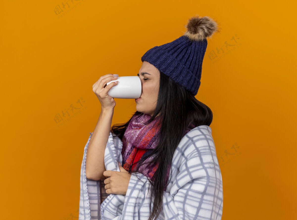 感觉愁眉苦脸的生病的年轻女人戴着冬天的帽子 围着格子呢围巾站在侧视图里喝着茶杯闭着眼睛孤立地站在橙色的墙上冬天皱眉市民