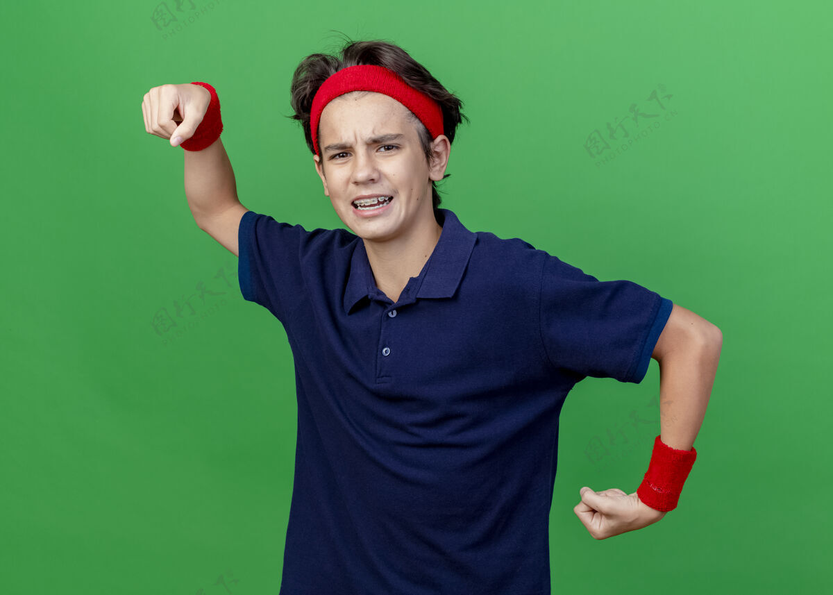 运动自信的年轻帅气的运动男孩戴着头带和护腕 戴着牙套 做着强烈的手势 看着绿色墙壁上孤立的前方姿势人衣服