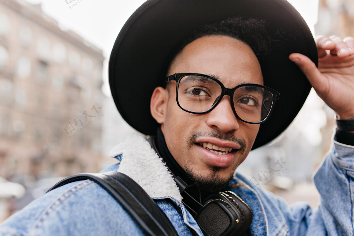 享受棕色眼睛的非洲男人抚摸着他的黑帽子的特写镜头欣喜若狂的混血男模走在街上的户外镜头情感年轻城市