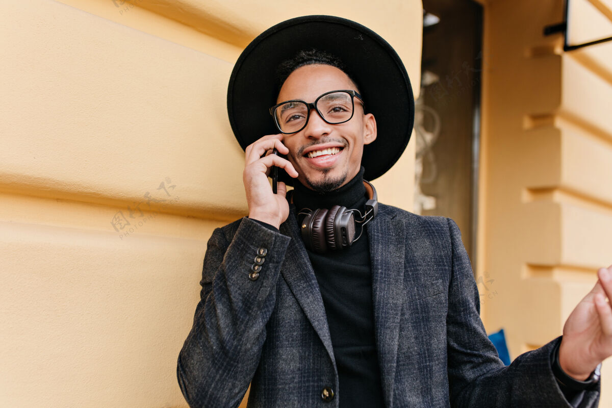 电话穿着羊毛夹克的快乐的非洲男人打电话给朋友的户外肖像戴帽子的快乐的黑人男人站在黄色大楼附近打电话快乐手机年轻