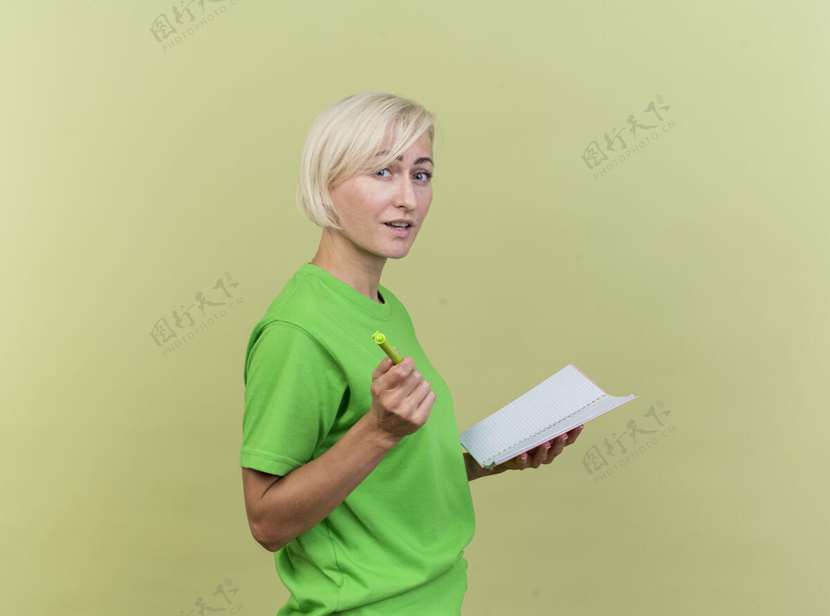 笔令人印象深刻的中年金发女人站在侧视图中拿着便签本指着前面 笔孤立在橄榄绿的墙上人姿势印象