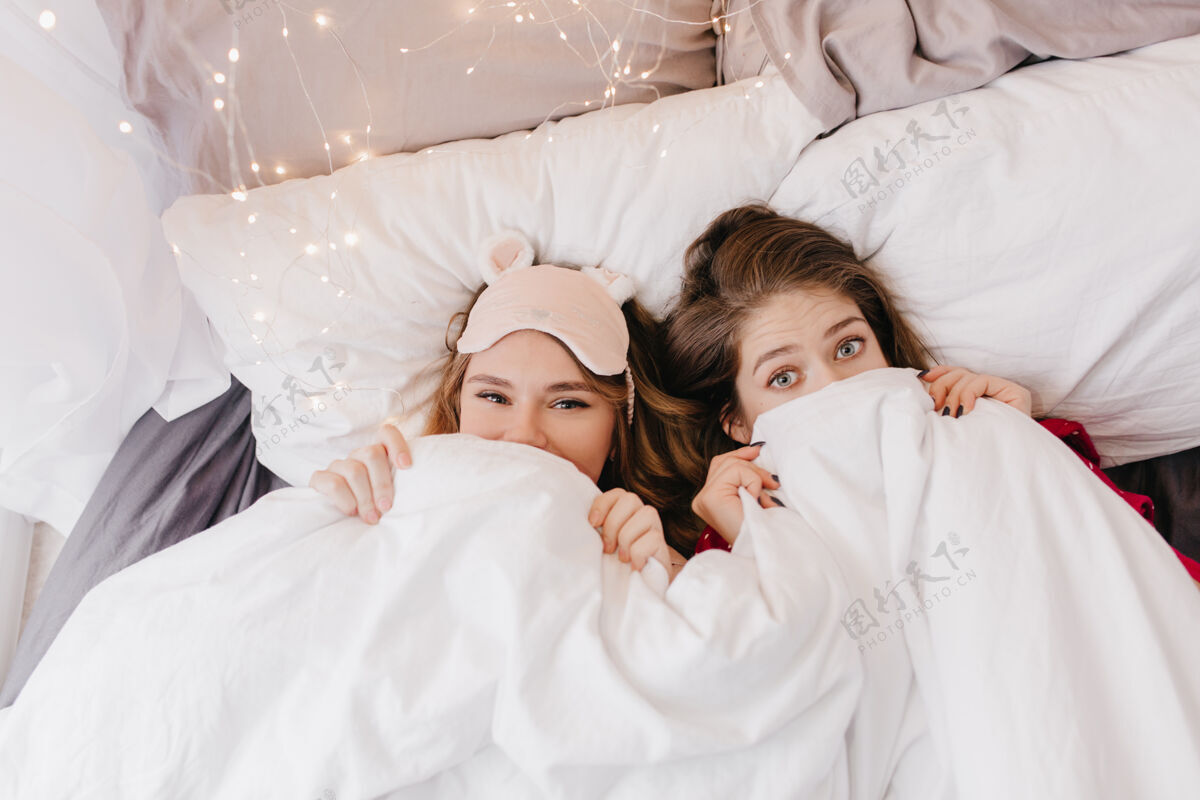枕头漂亮的金发女孩 戴着粉红色的睡衣 躲在毯子下面两个优雅的姐妹在早晨的照片中开玩笑的室内照片眼罩表情快乐