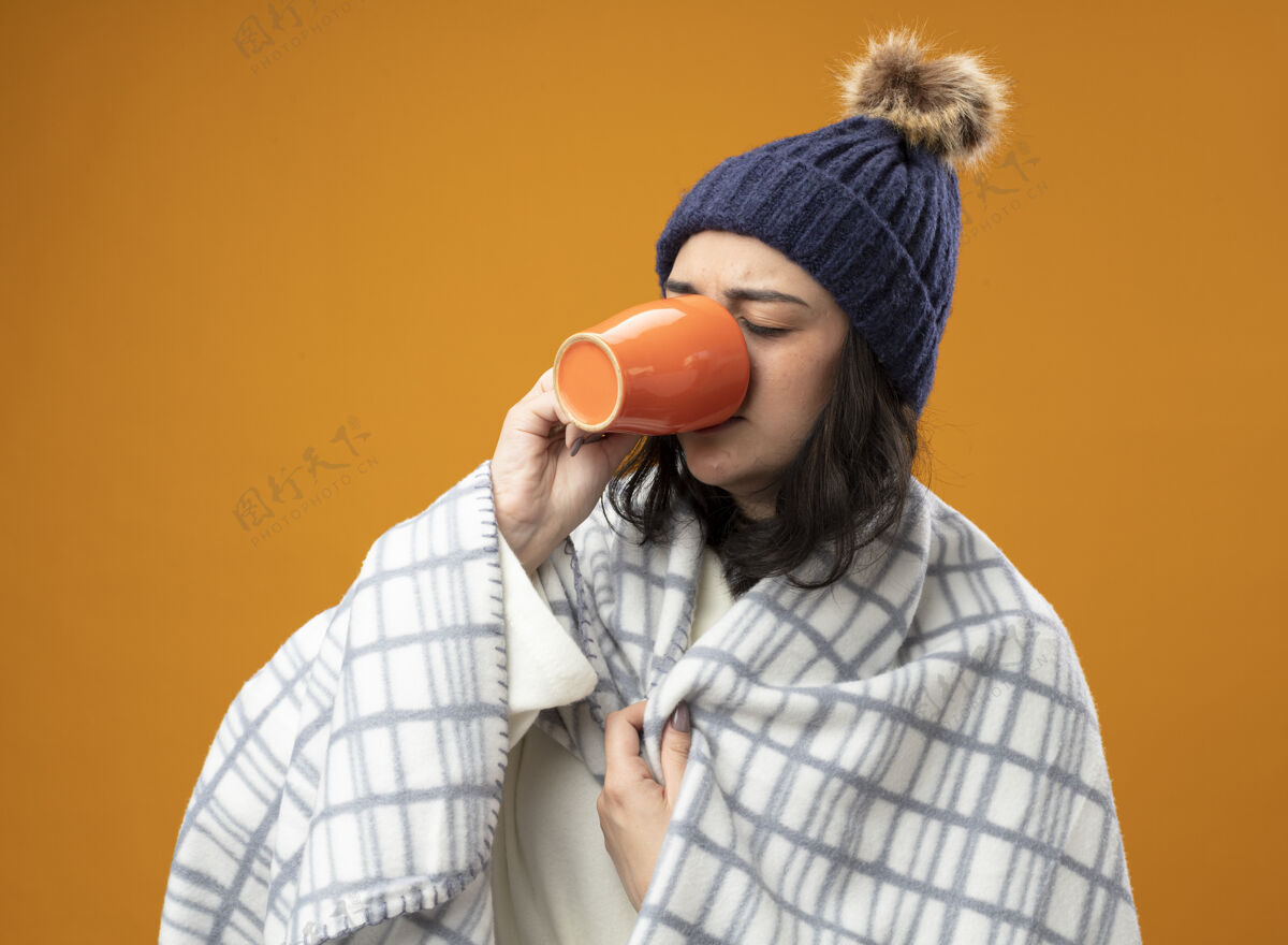 闭嘴愁眉不展的年轻病女人穿着长袍 戴着格子布裹着的冬帽 喝着一杯茶 紧闭着眼睛抓着格子布 孤零零地站在橙色的墙上抓住衣服姿势