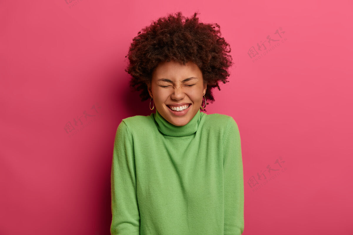 无忧无虑室内拍摄的迷人卷曲的年轻女子笑容灿烂 露出洁白的牙齿 笑出有趣的笑话 表达真诚的情感 在粉红色的墙上摆姿势人 情感 生活方式的概念人快乐墙