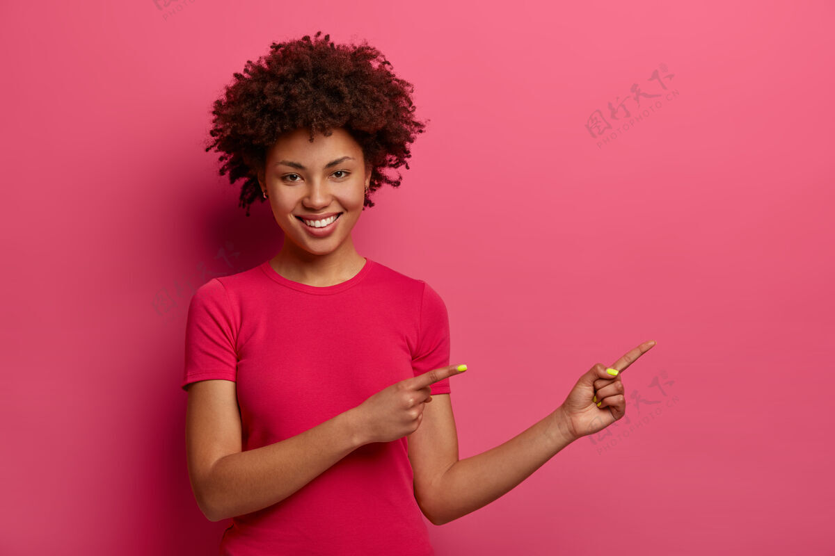 积极漂亮的卷发女人站在一边 推荐漂亮的产品 展示促销的地方 笑容满面 说试试这个 隔着粉色的墙给你的广告留个空间指向种族情绪