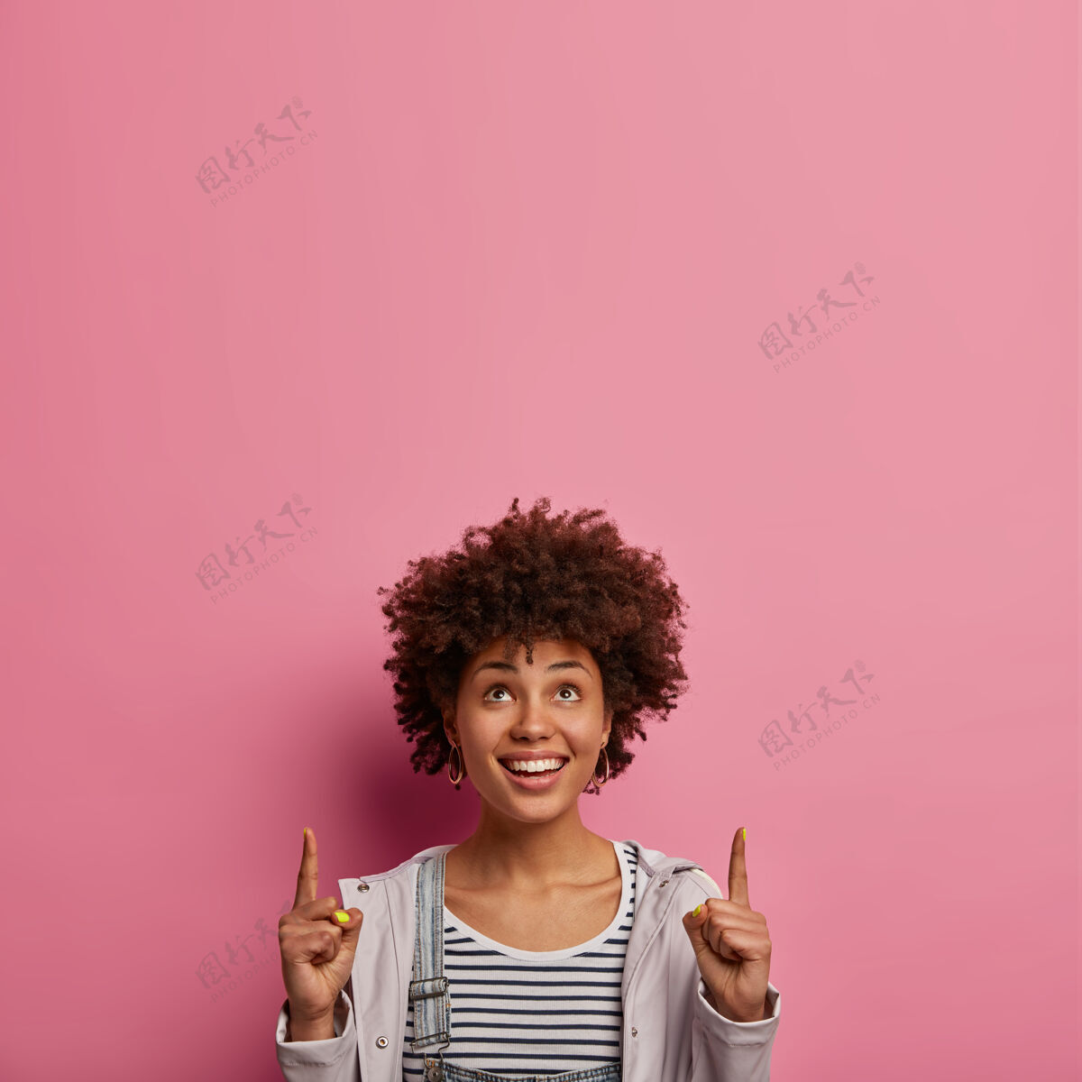 女士快乐的卷发提供令人难以置信的优惠 看起来很有趣 表示向上 有牙齿的微笑 高兴很好的销售 穿着随意 在玫瑰色的墙上摆姿势 建议参加有趣的活动无尾人非洲