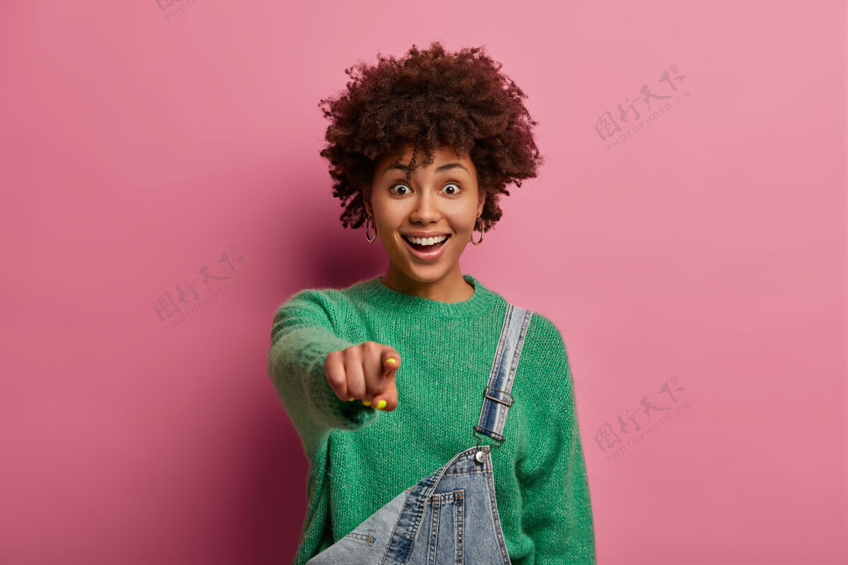 脸兴奋积极的女人有着非洲式的发型 食指直指你 看到一些很有趣的事情 穿着绿色毛衣和工作服 隔离在玫瑰色的粉彩墙上哇 太棒了人选择人