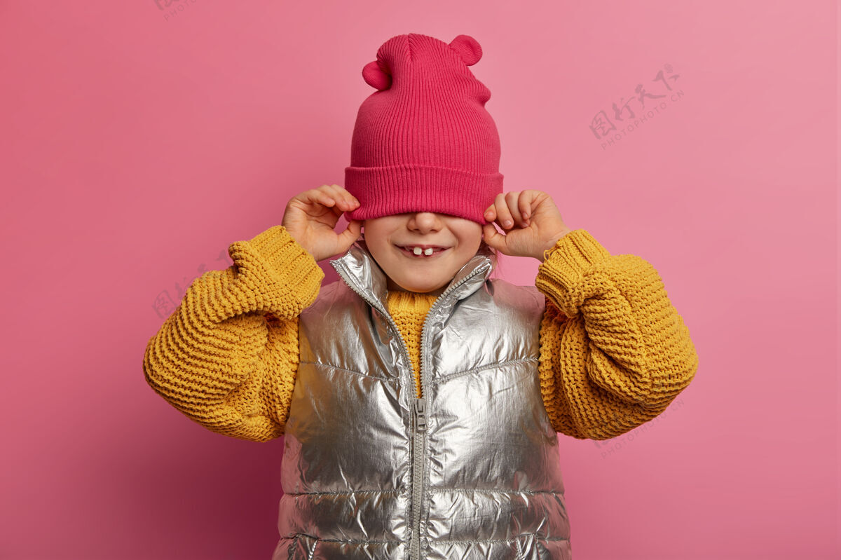 乐观可爱顽皮的小孩戴着帽子掩面 穿着暖和的毛衣和马甲 享受童年 在室内对着柔和的玫瑰色墙壁摆姿势 两颗牙齿突出休闲风格快乐情感概念室内孩子欧洲