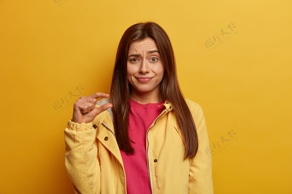 照片一张年轻黑发女子的照片 她用手指展示着非常小的身躯 展示着微小的身躯 讲述着降价和微薄的薪水 困惑地扬起眉毛 穿着黄色夹克钱包衣服无领
