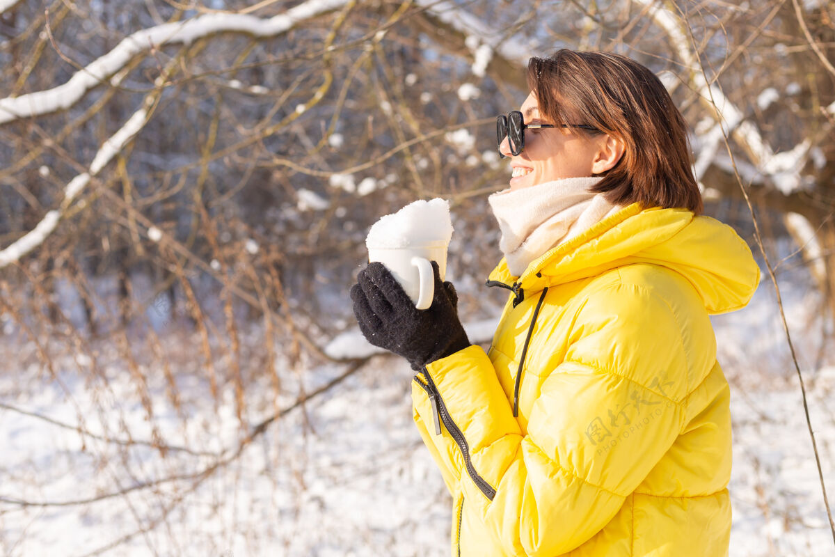 可爱美丽的年轻开朗的女子在雪景的冬日森林里戴着墨镜拿着装满雪的杯子玩得开心霜冻公园季节
