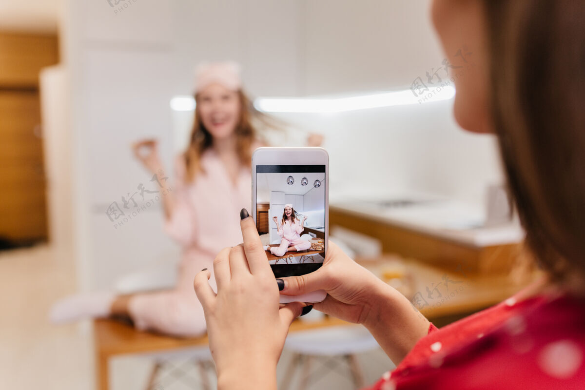 成人快乐的白人女孩吃着比萨饼 玩着她的头发黑发女人拿着智能手机 在厨房里给朋友拍照 室内光线明亮快乐肖像朋友