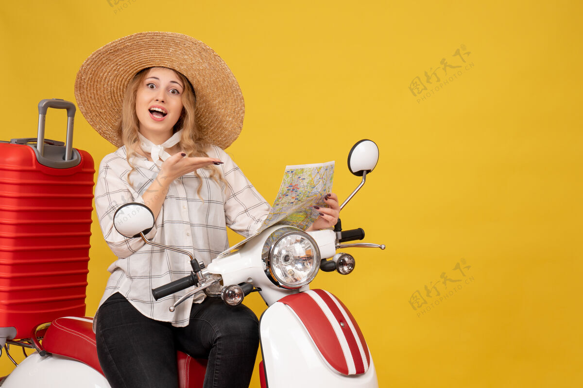 帽子戴着帽子坐在摩托车上的年轻女子的俯视图 黄色的脸上带着微笑的表情指着地图年轻女子摩托车面部