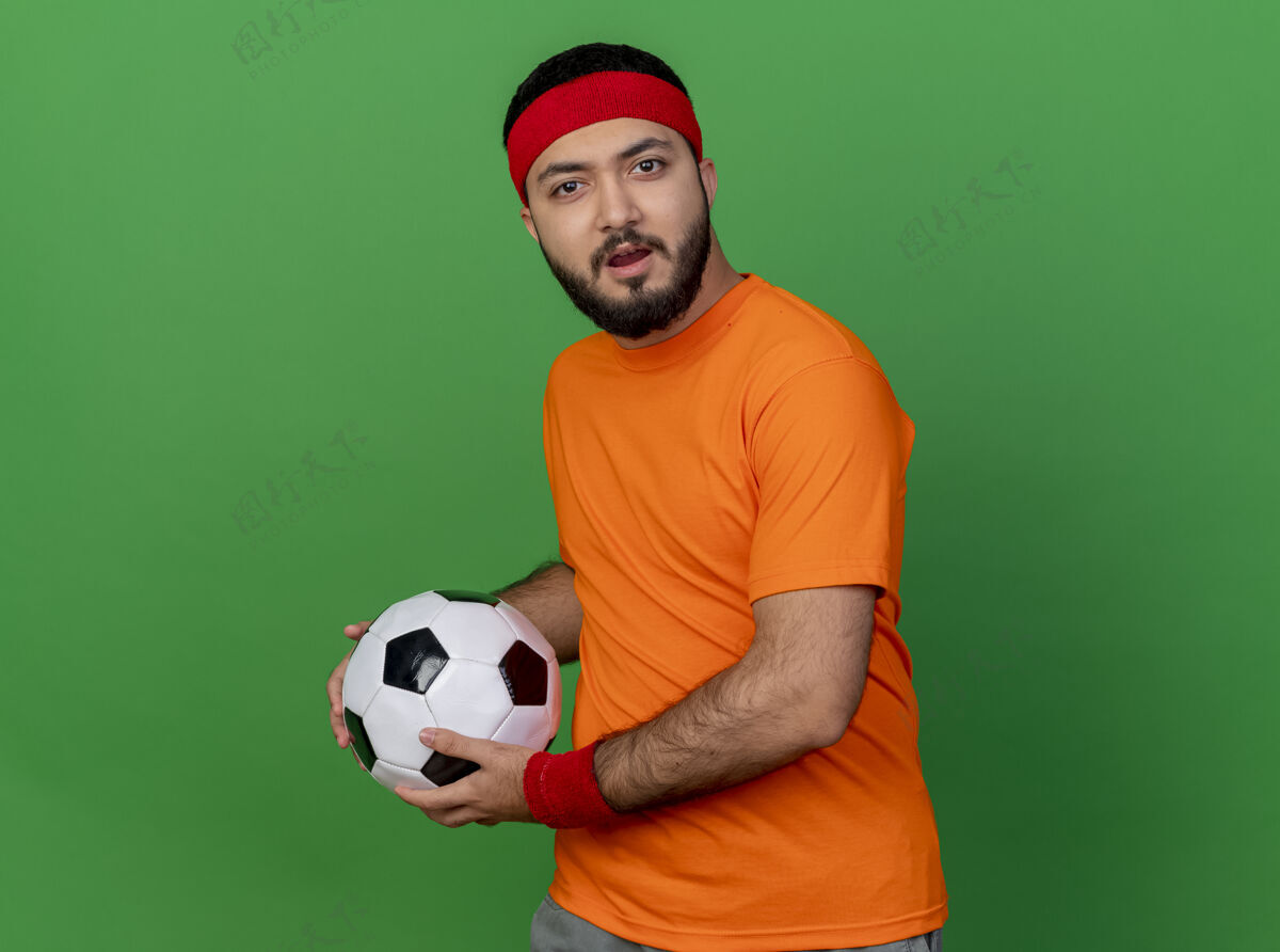 困惑困惑的年轻运动男子戴着头带和手环拿着球孤立的绿色背景球运动头带