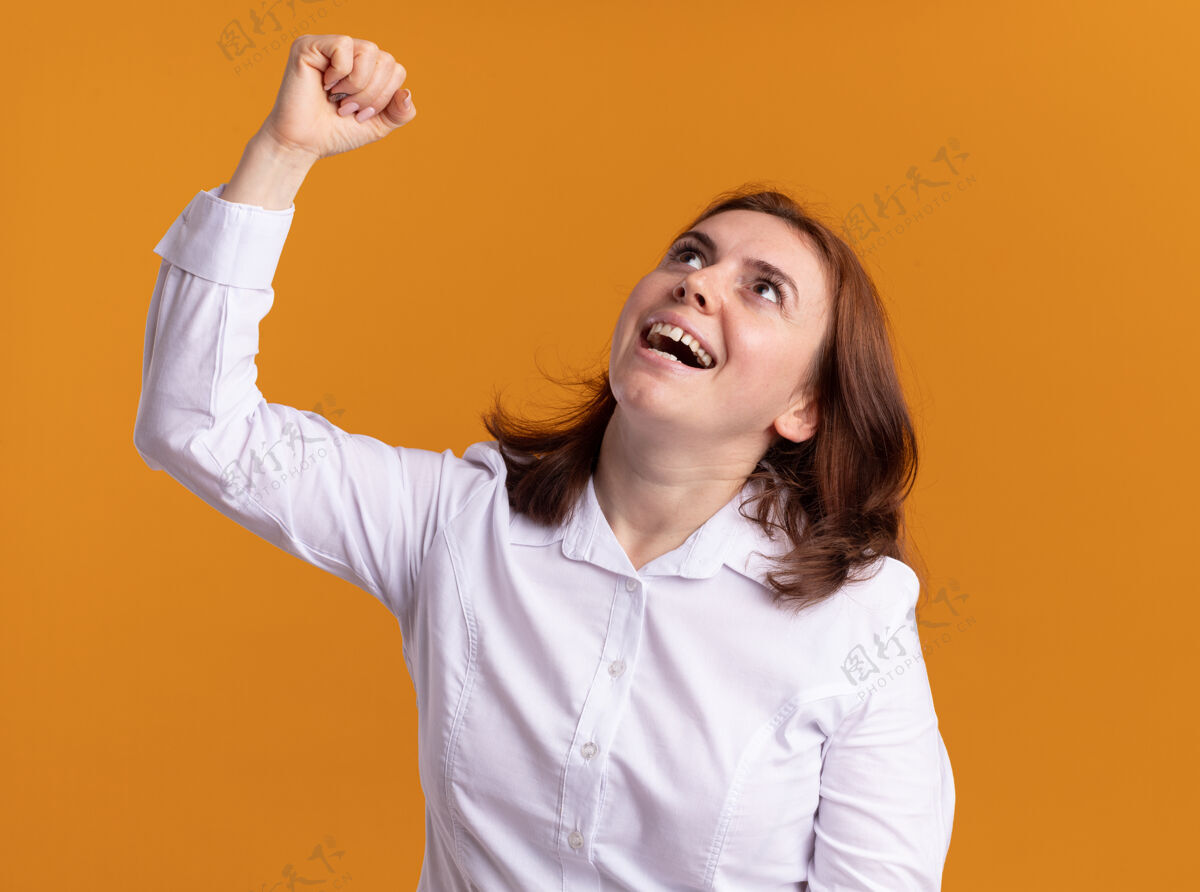 快乐身穿白衬衫的年轻女子高兴地抬起头 兴奋地举起拳头 像一个站在橘色墙壁上的胜利者赢家喜欢年轻