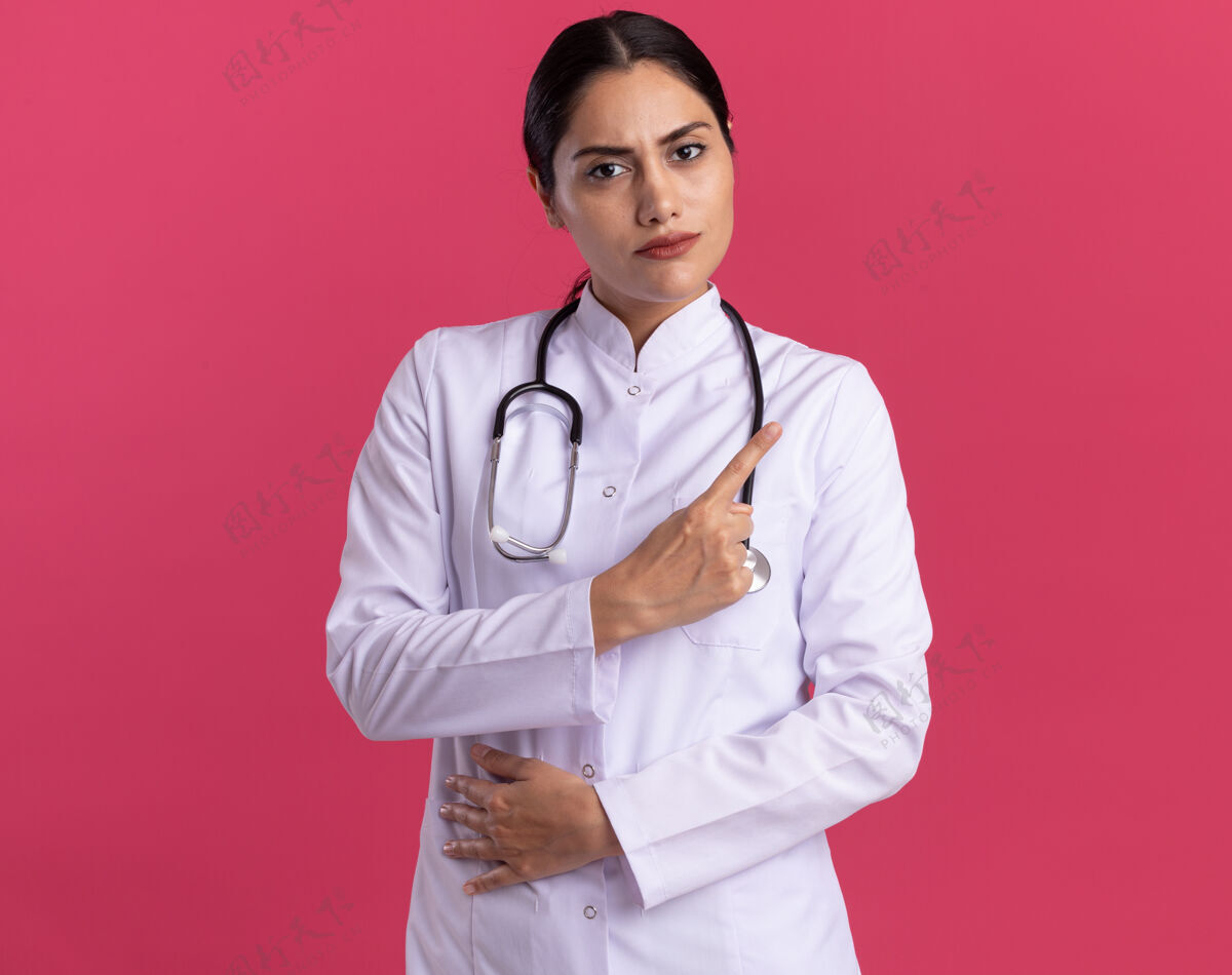 女人身穿医用外套的年轻女医生带着听诊器看着前方 严肃的脸上露出食指警告站在粉红色的墙上警告医生严重