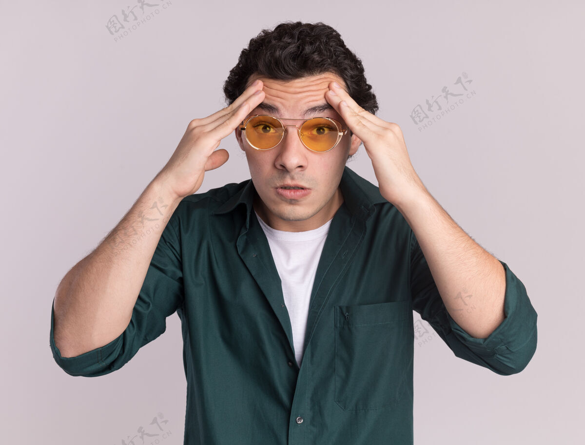 衬衫一个身穿绿衬衫 戴眼镜的年轻人站在白墙上 摸着头看着前面 脸上带着头痛的恼怒表情年轻头表情
