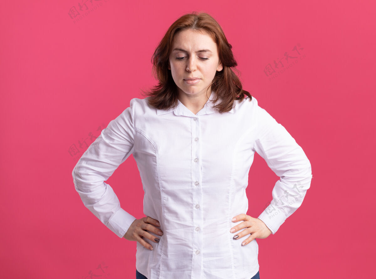 手臂身穿白衬衫的年轻女子 面带严肃的表情 双臂放在臀部 站在粉红色的墙上站立女人严肃