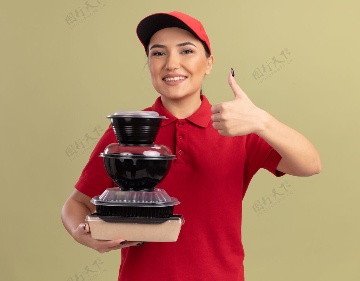 抱着身穿红色制服 头戴鸭舌帽的年轻女送货员站在绿色的墙上 手里拿着一叠食品包 面带微笑 兴高采烈地竖起大拇指看着前面欢呼帽子食物