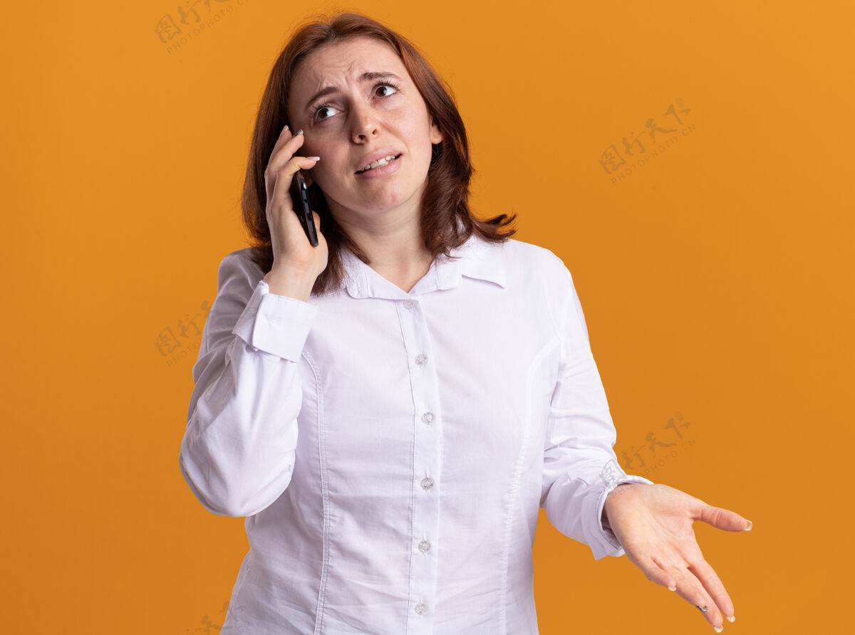女人身穿白衬衫的年轻女子站在橘色的墙上讲手机 感到困惑年轻电话衬衫