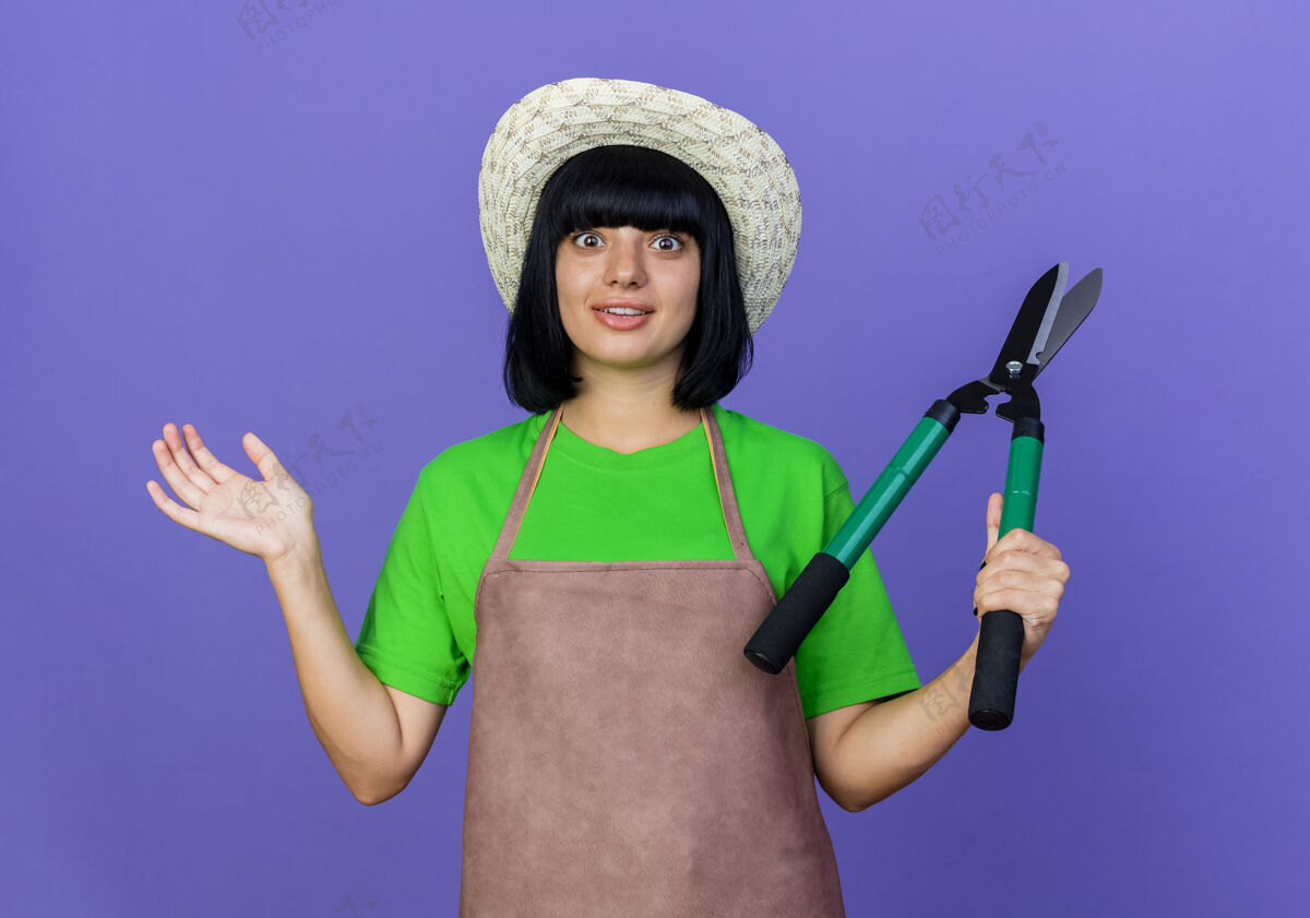 紫色穿着制服的年轻女园丁惊讶地戴着园艺帽拿着剪刀花园复制帽子