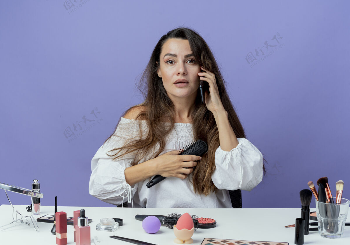 女人惊讶美丽的女孩坐在桌子旁 拿着化妆工具 拿着梳子 在紫色的墙上讲电话化妆坐着发夹