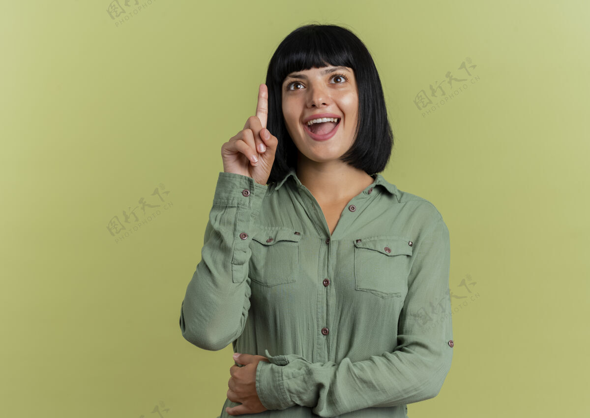 尖兴奋的年轻黑发高加索妇女指出孤立的橄榄绿背景与复制空间黑发背景女人
