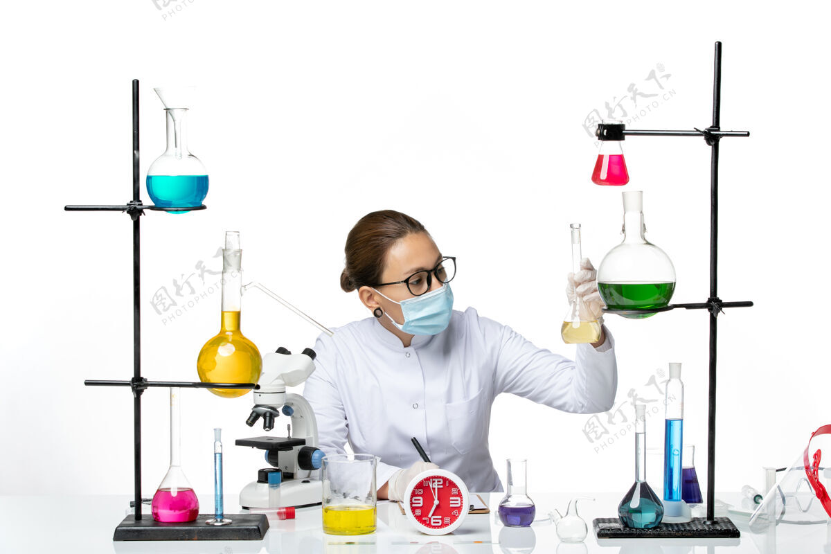 医学前视图：穿着白色医疗服的女化学家戴着面具在白色背景上写笔记化学家实验室病毒冠状病毒飞溅研究喷溅烧杯