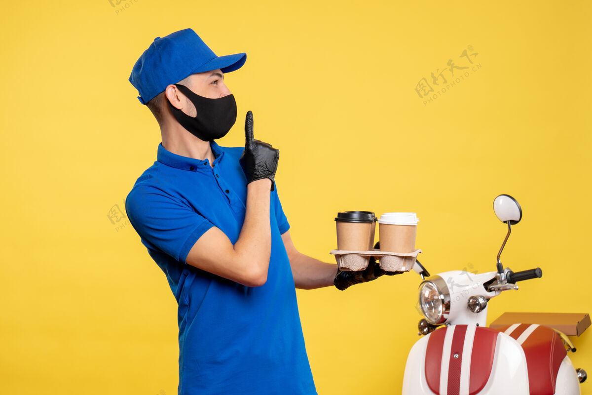 持有正面图身穿蓝色制服 戴着面具的男信使拿着咖啡 穿着黄色工作服 工作服 covid-送货大流行专业帽子蓝色