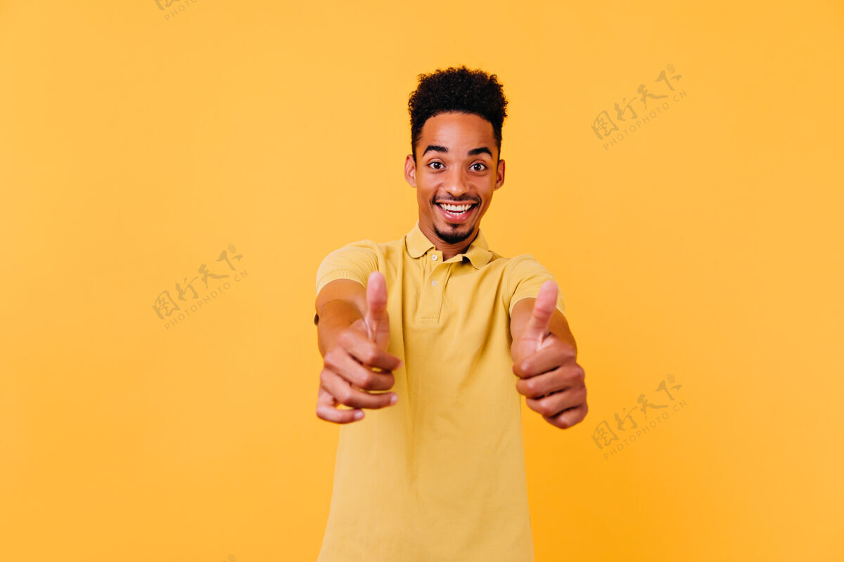 配饰优雅的黑人男孩露出惊讶的微笑 竖起大拇指幽默的非洲男人的室内照片 滑稽的发型幸福舞蹈寒冷