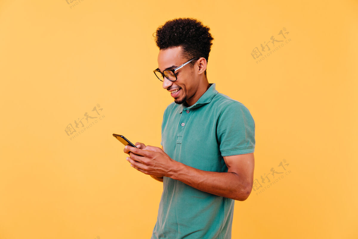 黄色感兴趣的黑人男子带着愉快的微笑看着手机屏幕戴眼镜的英俊非洲男孩阅读留言的室内镜头设备黑色沟通